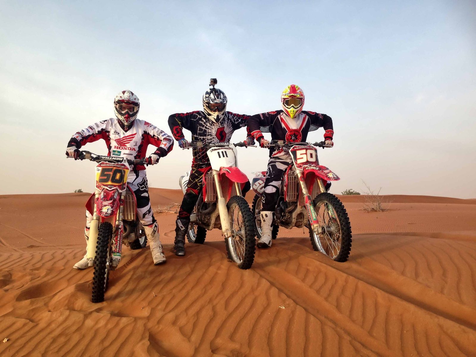 Motocross in Dubai: three riders standing with their bikes in the orange sand of the desert