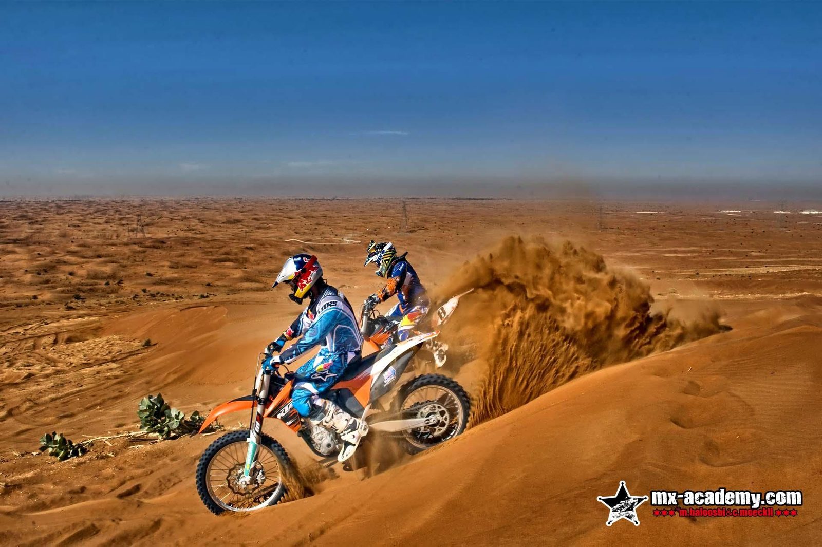 Motocross in Dubai: two riders in the desert dunes, sand spraying up behind them under a blue sky